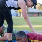 A photo of Kate Rowen bending over to help a man doing a plank.